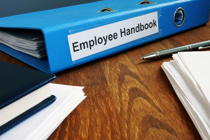 Blue binder labeled "Employee Handbook" on a wooden desk, surrounded by loose papers and a pen, conveying an organized and professional setting.